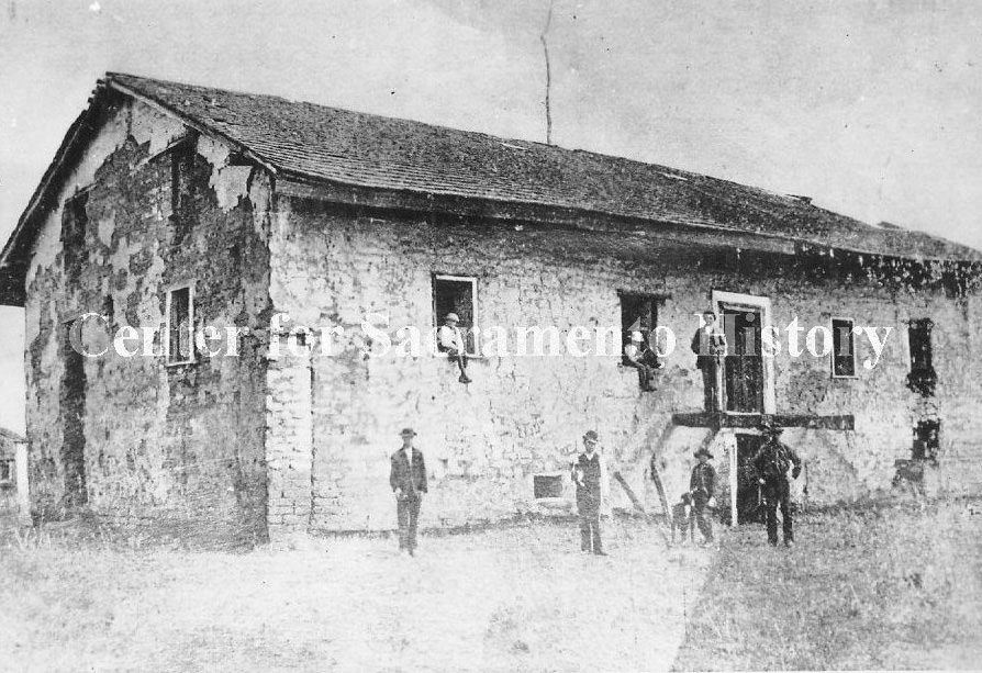 Sutter's Fort prior to being rebuilt with just the central building left standing. Several men and a dog are sitting in windows and doorways and standing next to the buiding, 1867