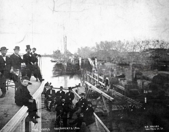 The Levee Sacramento 1866. The photo shows boats, railroad tracks with train, and men seated on boat in foreground.