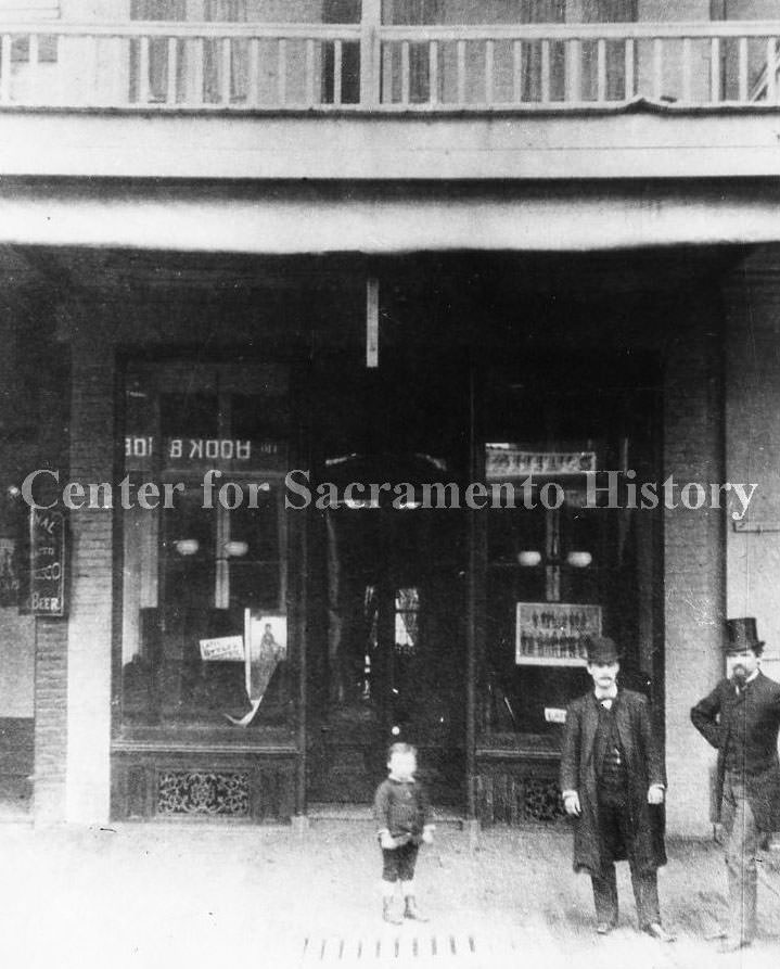 Store front on J Street with a men and a small boy posing in front of the store, 1868