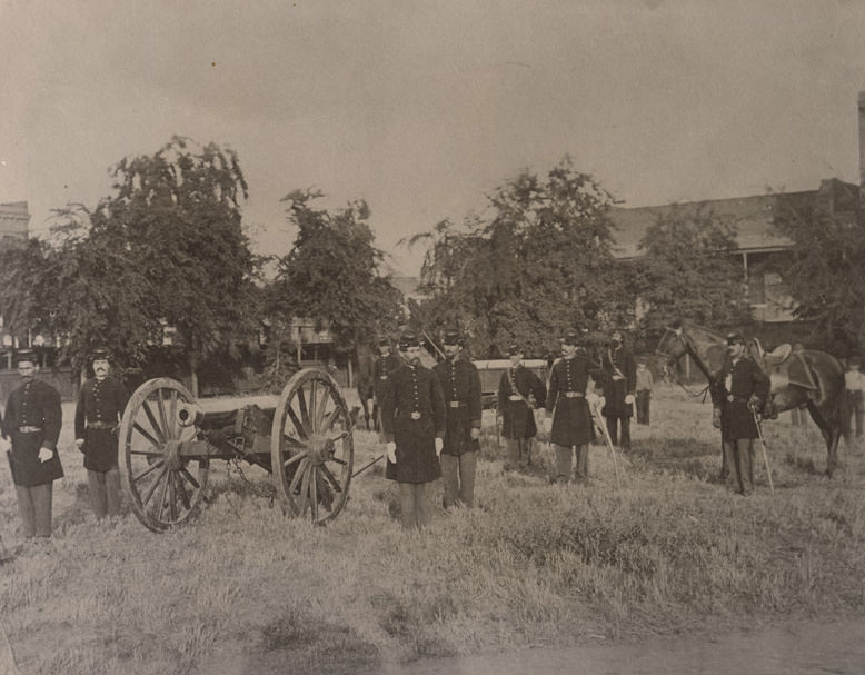 Nine members of the California Milita Batter B. An 1841 Howitzer Cannon at left. The men stand in grassy area, bldgs. at rear, Sacramento City Plaza, 1865