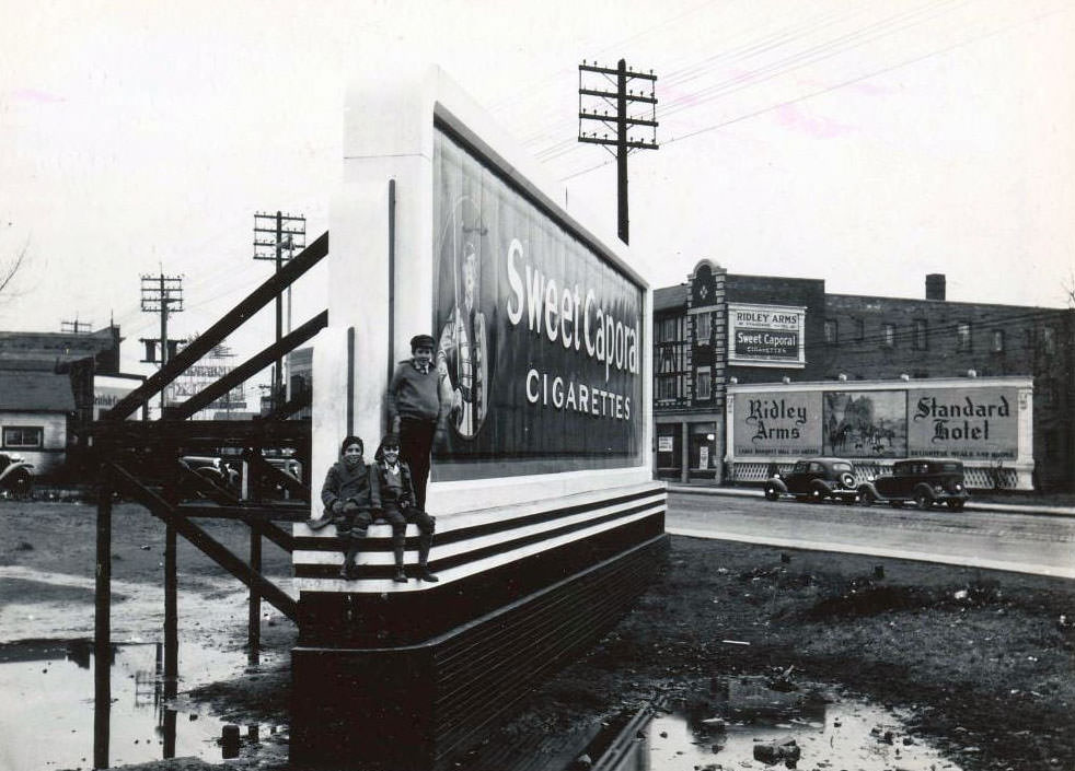 Three young boys are posing on the base of the board. The billboard is opposite the Ridley Arms, a Standard Hotel, at 2301 Danforth Avenue, 1936