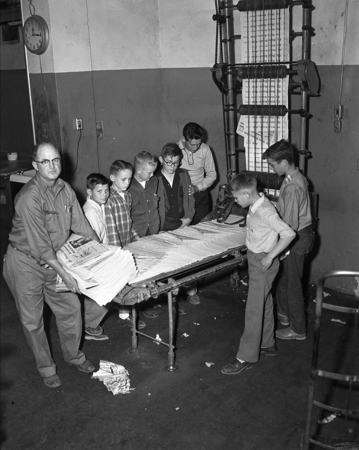 Abilene Reporter News boys and a man standing around a newspaper printer, looking at the stack of newspapers on the machine, 1955