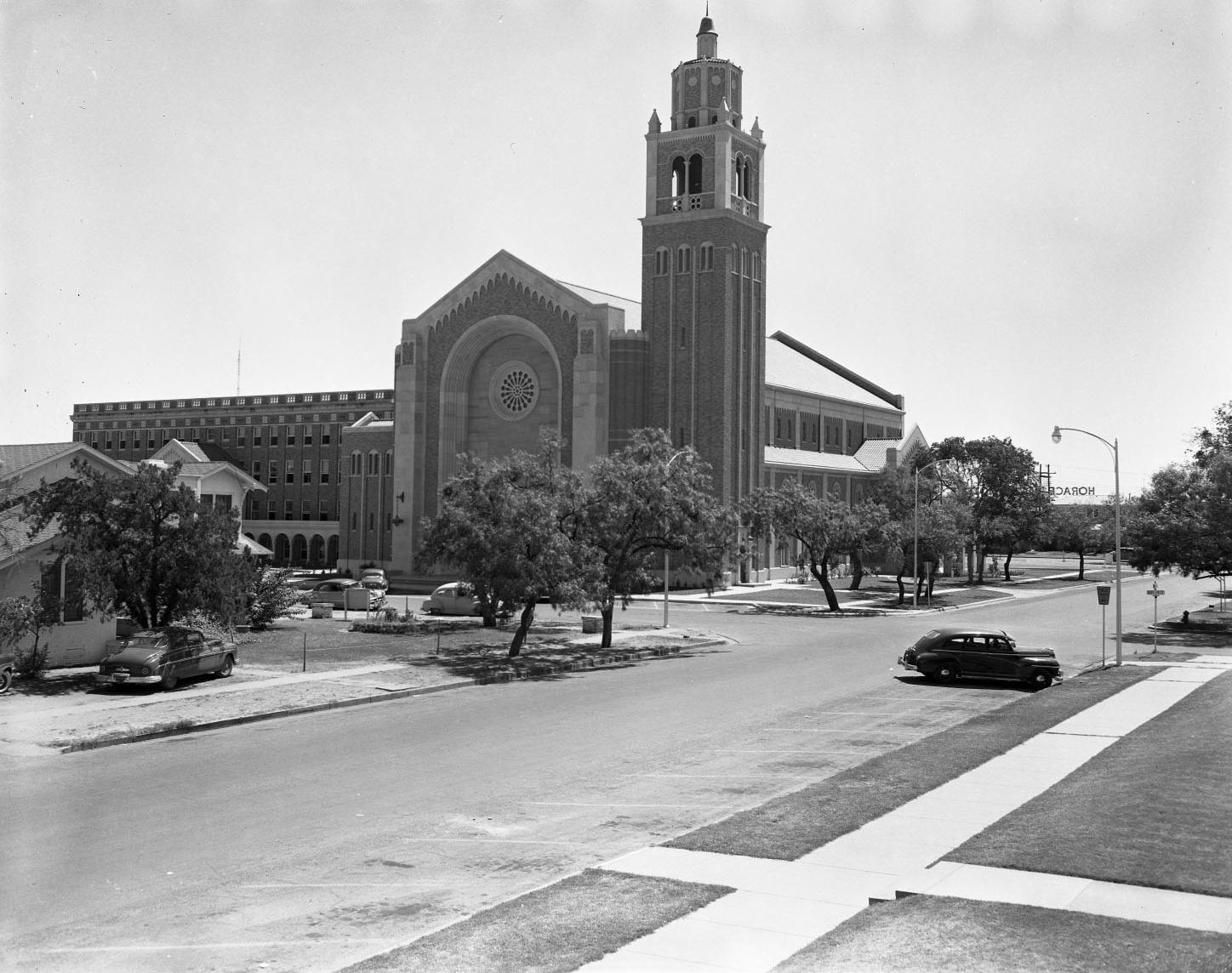 Exterior view of First Baptist Church in Abilene, 1958. The church has Gothic style architecture and a large bell tower. There are cars parked in the street outside the church.