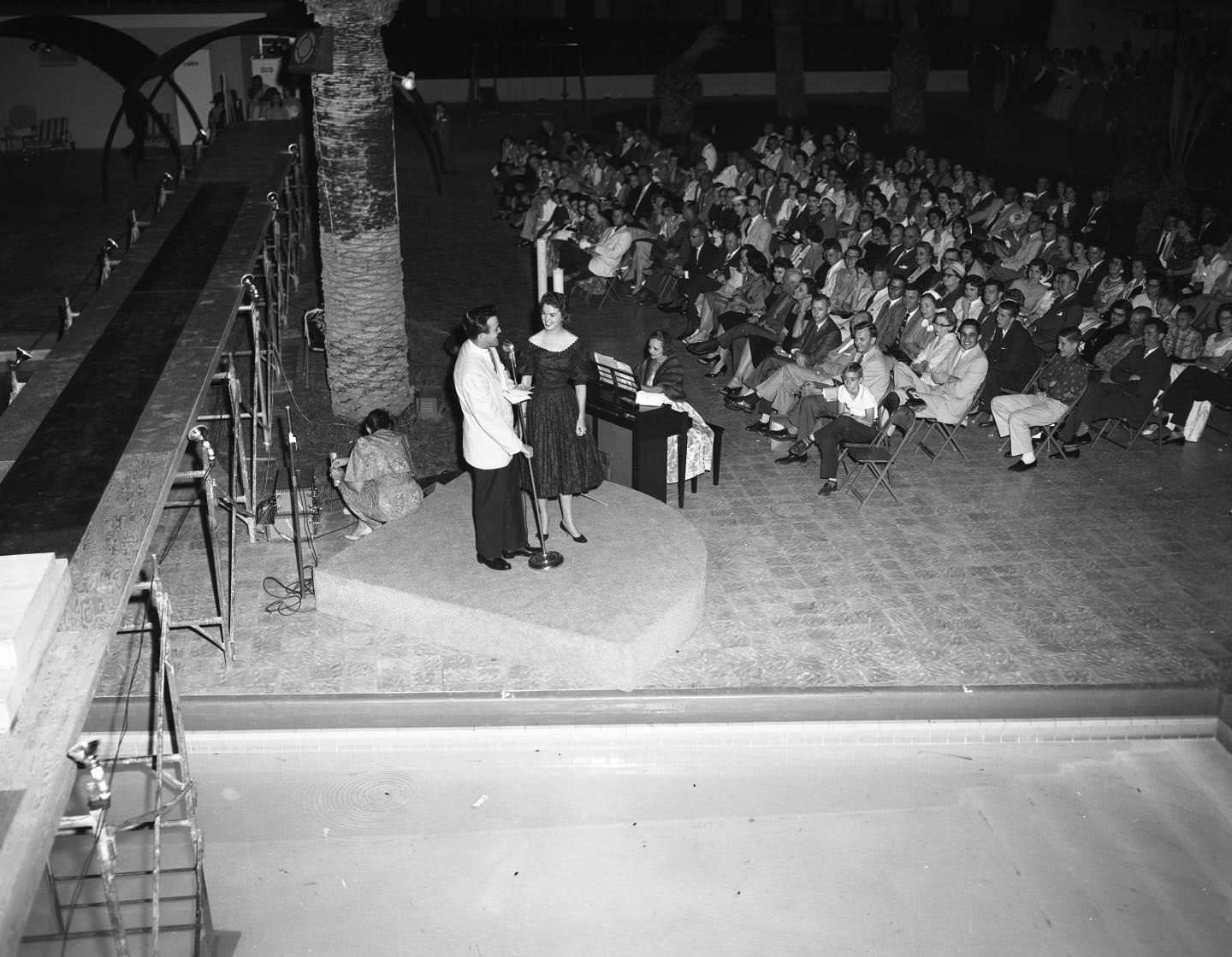 A crowd at a beauty pageant contest. They are all sitting while a man and a woman appear to sing on a small stage, 1958