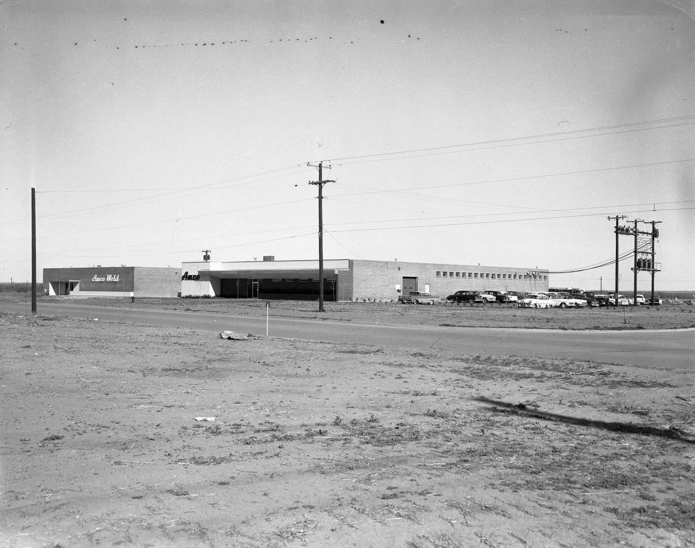 Two Amco buildings on West N. 1st Street in Abilene, 1958. The buildings are viewed from across a street, and there are cars parked beside them and power lines around them.
