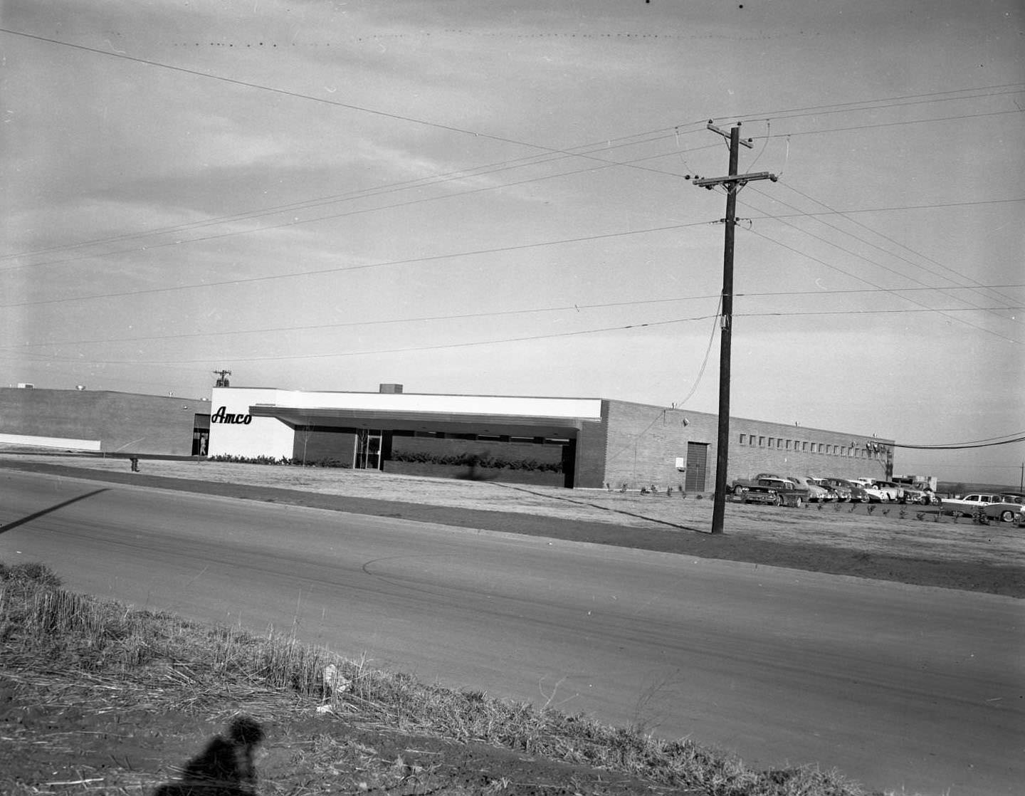 AMCO Manufacturing, 1958. To the side of the building, there are several cars parked in a parking lot. In the foreground, there is a street.
