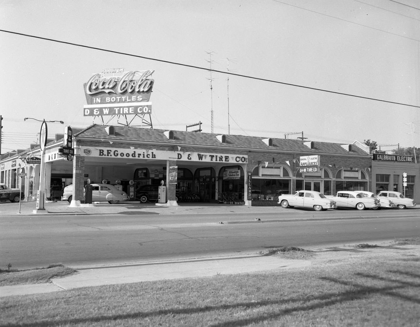 The D and W Tire Company, 1956. There are a few cars parked in front of the store and one car appears to be getting serviced. Galbraith Electric is next to the store.