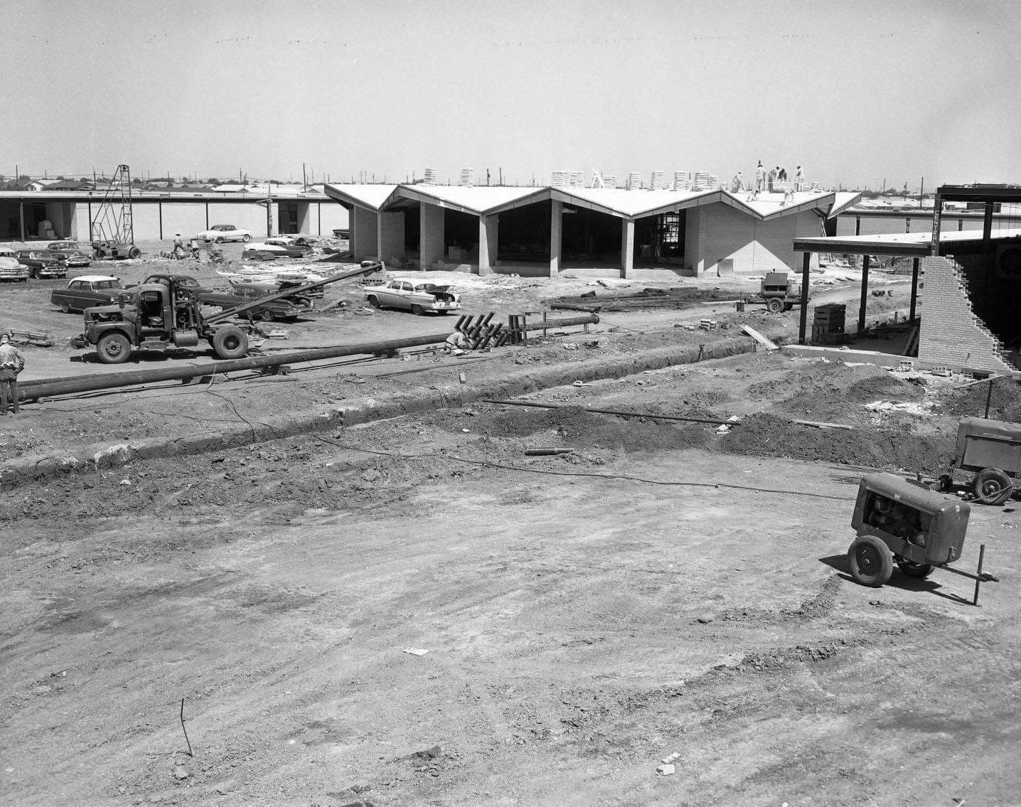 The construction of Cooper High School, 1955. Several buildings are all partially built. There are various pieces of construction machinery around the buildings. Several men stand on top of one of the buildings.
