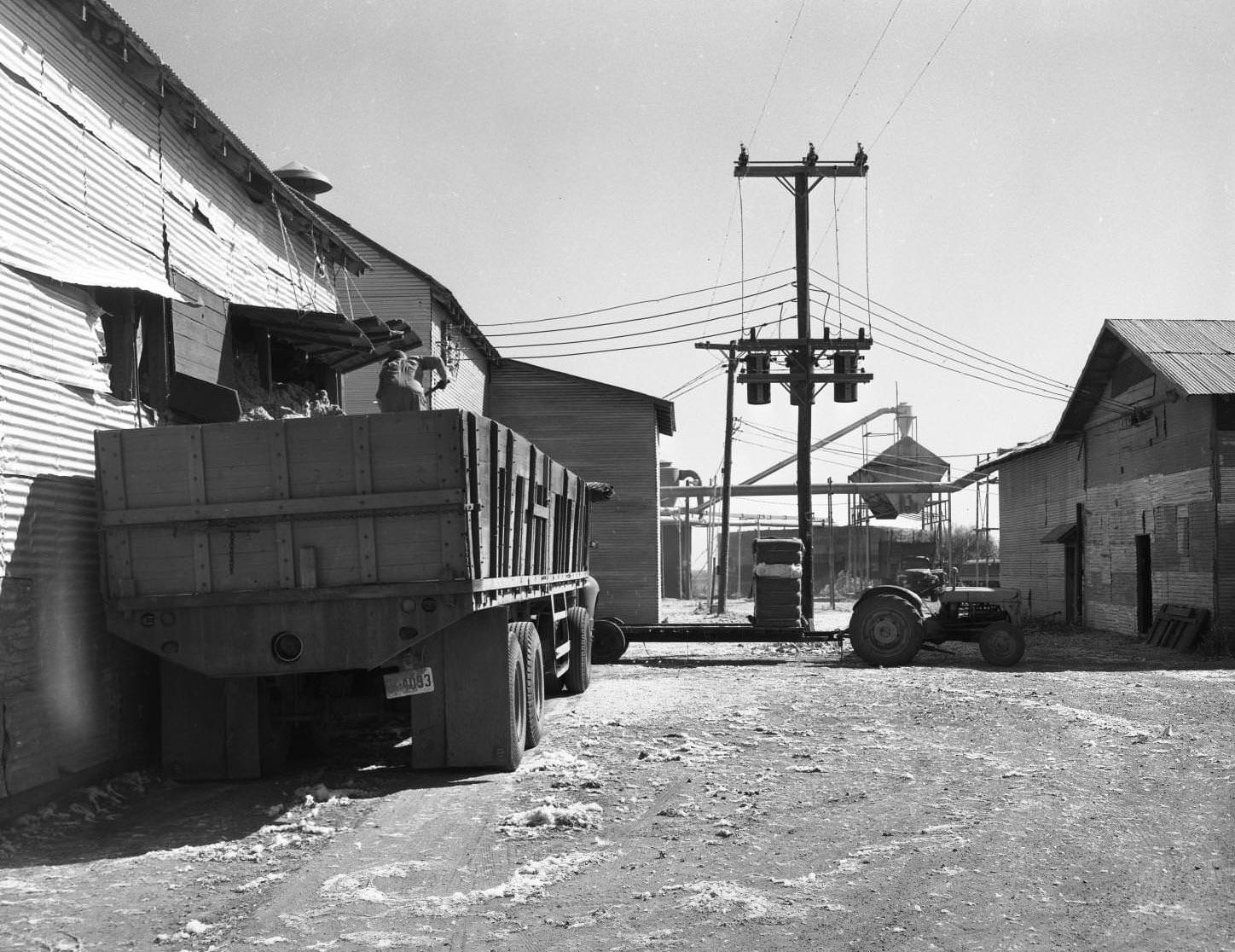 A cotton gin, 1958. There is a truck and trailer being loaded with cotton by a man. There are several buildings and machines with a tractor and power lines between them.