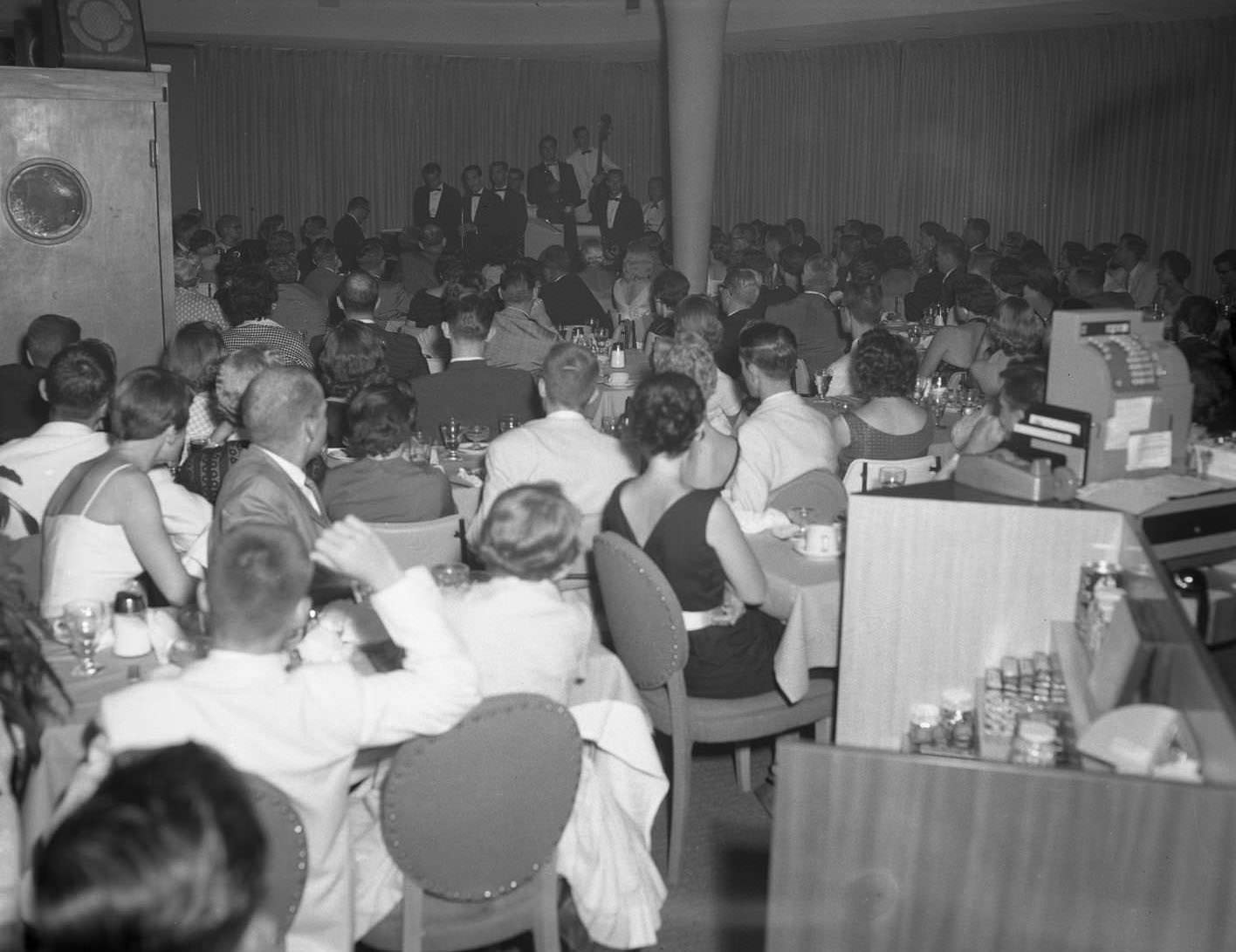 A show at the Sands Hotel in Abilene, 1957. The Crew Cuts quartet is singing on stage at the back of a room with an audience sitting around tables.