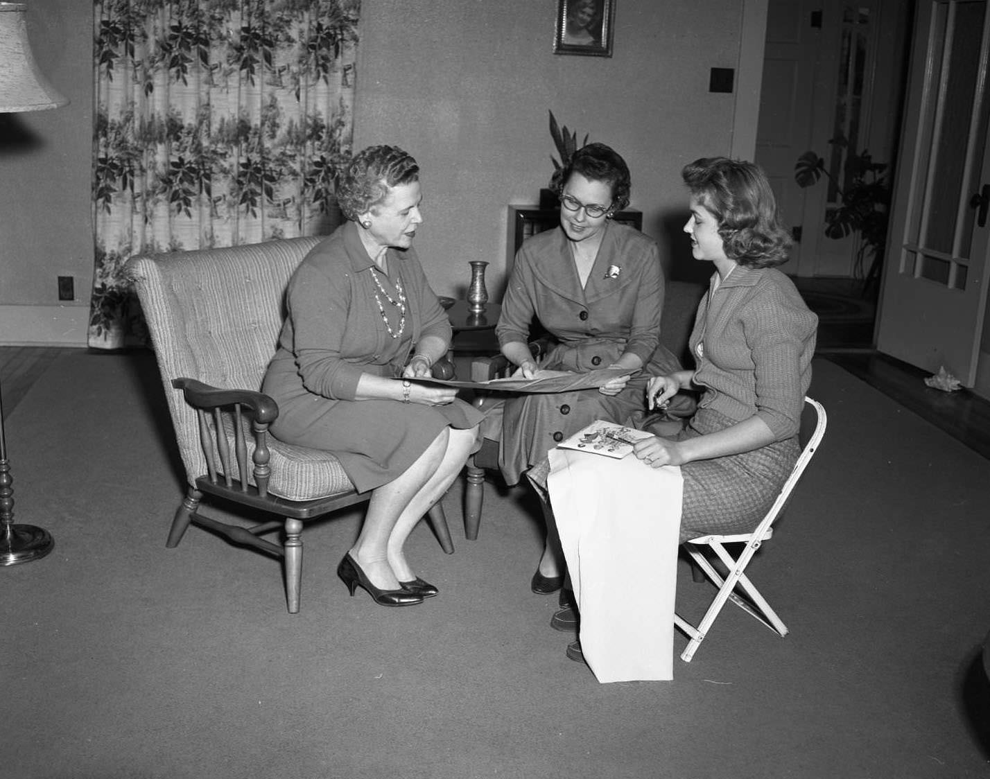 Three women in skirts sitting together looking at what appears to be a large piece of paper, 1958. The woman on the right appears to have a small paintbrush in her hand.