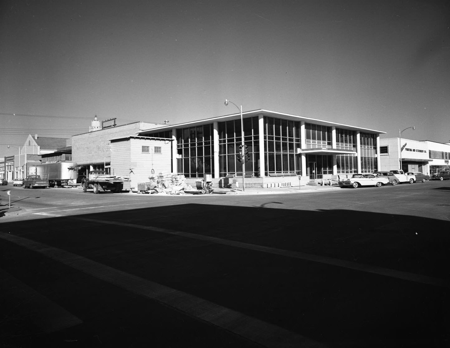 The Abilene Public Library, 1957. The building is on the corner of Cedar and North 2nd Street. There are other buildings on each side of the library, cars parked on the street, and light posts.