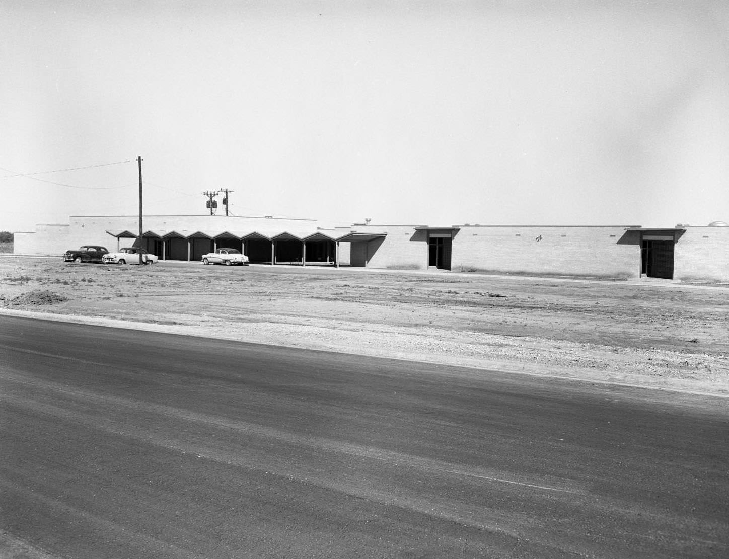 Elementary School Under Construction, 1958. In the front of the building, there is an awning and three cars parked beside it.