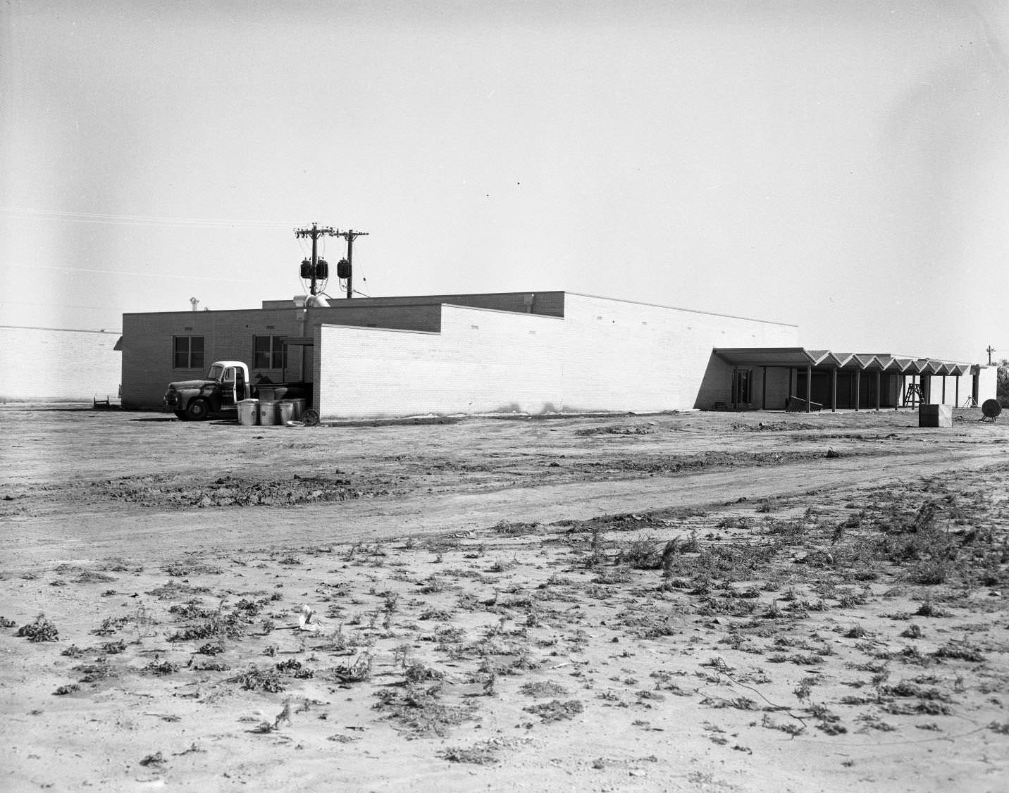 Elementary School Under Construction, 1958. The building has awnings, and there are vehicles parked beside it. It is in a bare field with power lines around it.