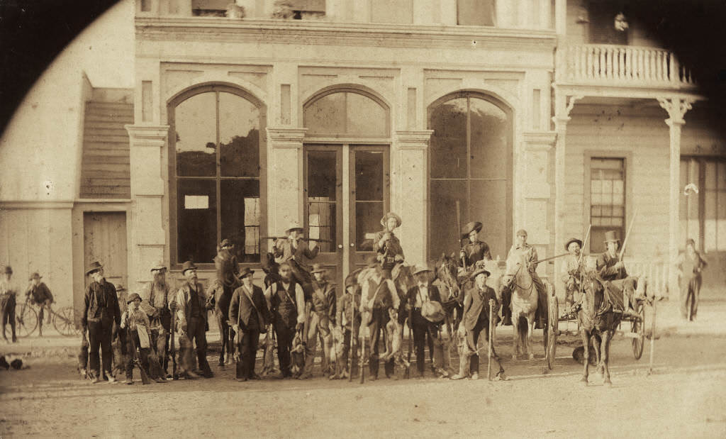 Rabbit hunting party standing in front of three large glass windows of the barber shop owned by Frank Ey on West Center Street (now Lincoln Ave.), Anaheim, 1893