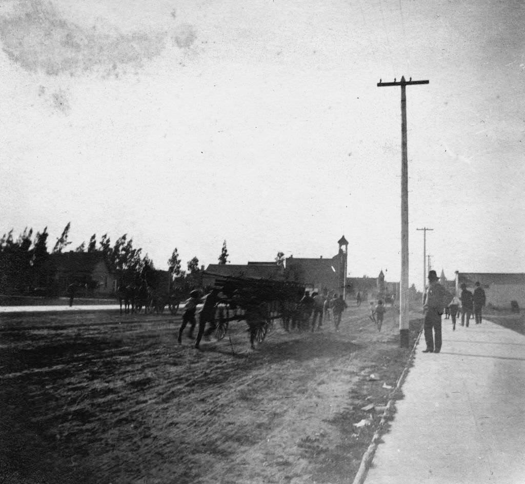 Anaheim Volunteer Fire Department with several men pushing and pulling a four-wheeled ladder wagon down Center Street (now Lincoln Ave.), Anaheim, 1895