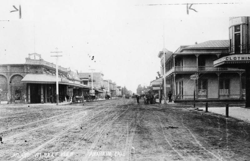View of an unpaved street in Anaheim in 1890, with shops and stores on either side and carriages parked at curbs. A streetcar is seen on tracks in the middle of the street.