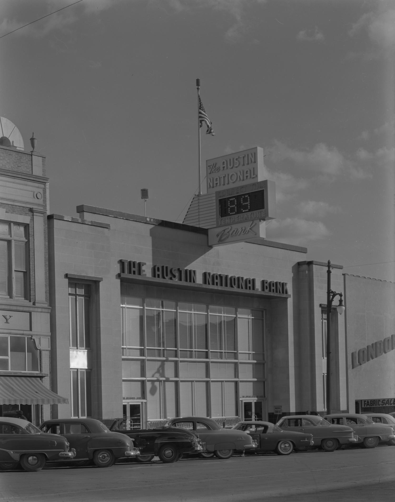 A street-side view of the Austin National Bank with a row of cars parked on the street in front of the building, 1954