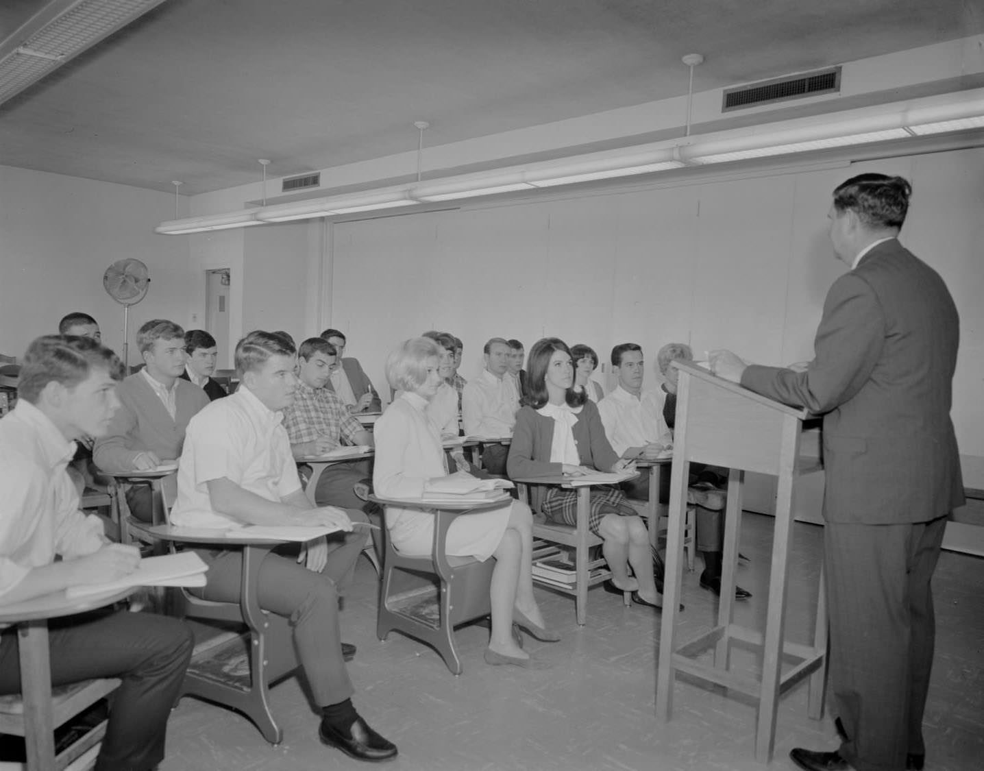 Students sit in desks looking at a man, presumably a lecturer, who is standing behind a wooden podium and speaking in an unknown room at St. Edwards University, 1966