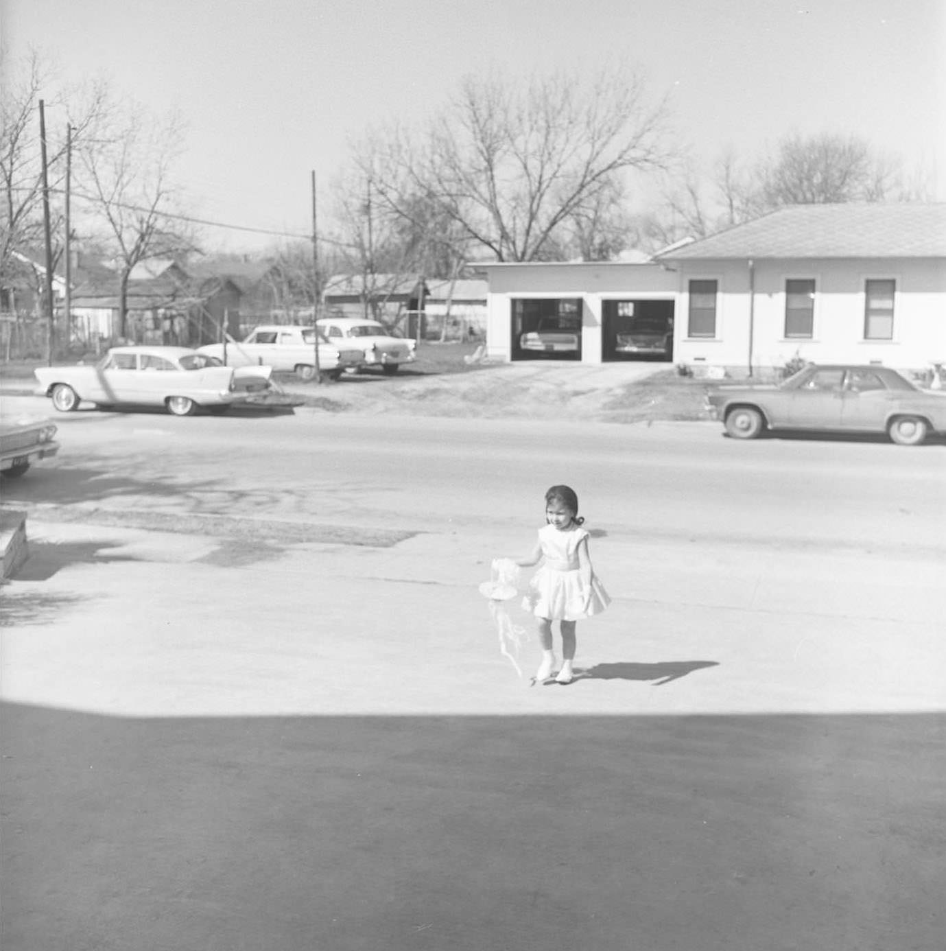Child of couple stands with bow in a driveway, 1965