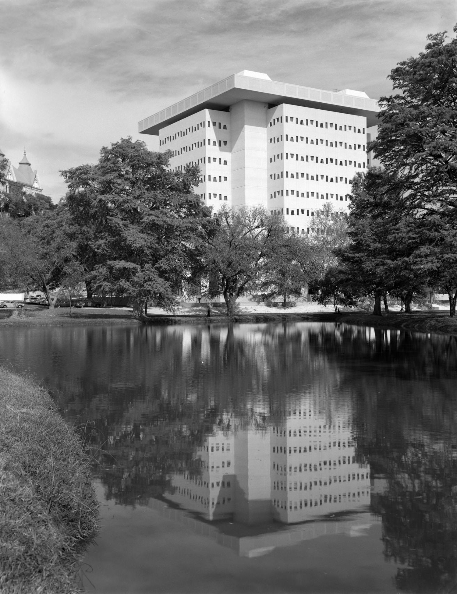A pond surrounded by trees near a tall, postwar building, 1969