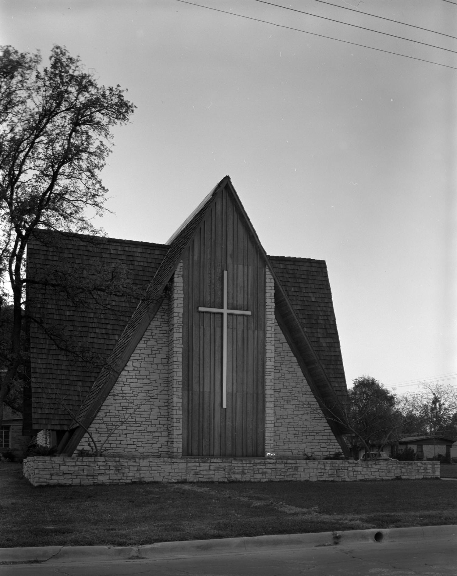 A church made from stone, with large, sloping roofs. On the building's front is a large cross, 1961
