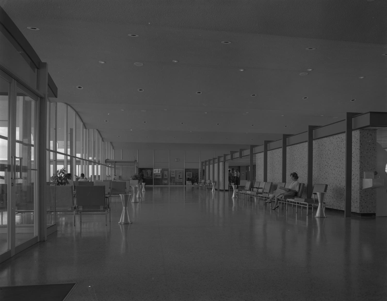 Interior of the Austin Municipal Airport where an open corridor can be seen with seating areas to the left and right, 1961