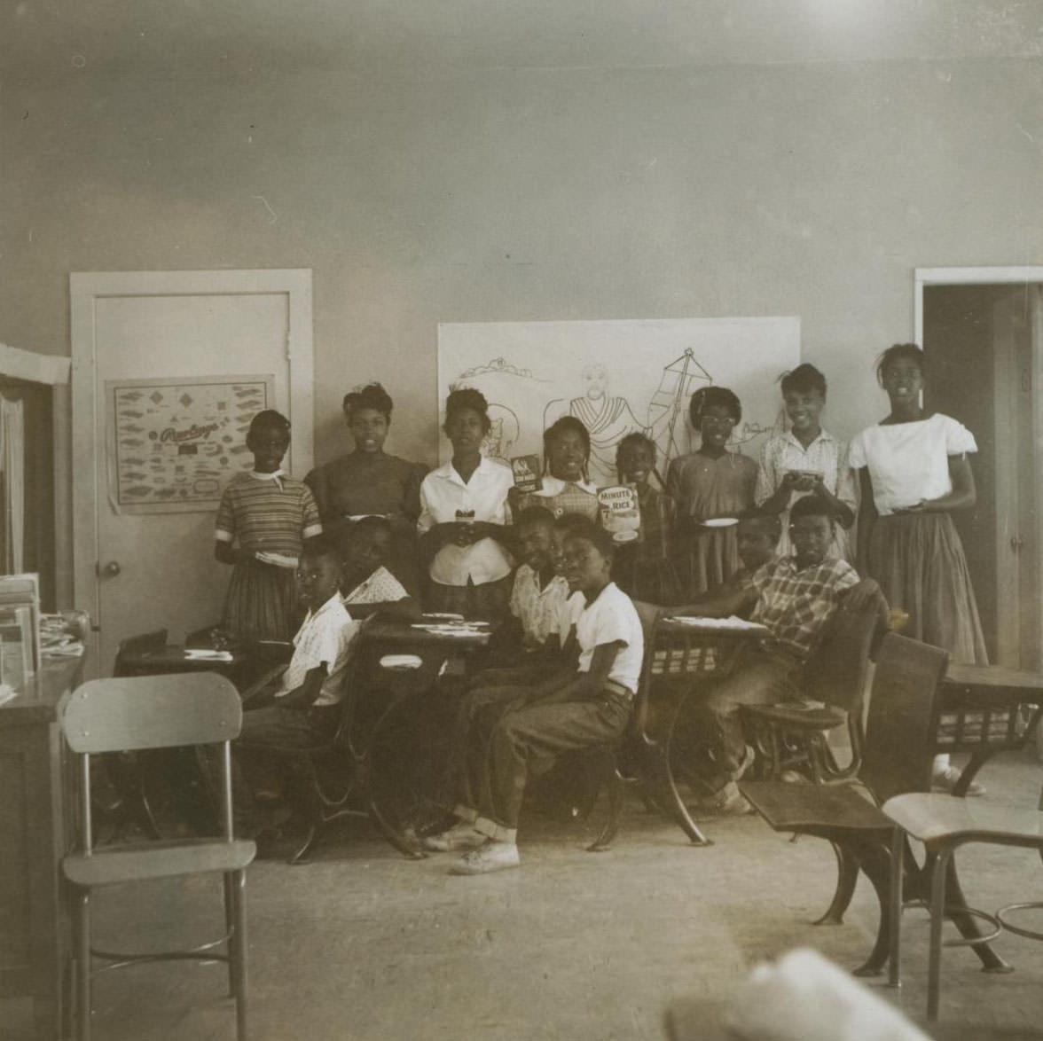 A group of students in a classroom, 1962 Boys sit at desks and girls stand against a wall behind them.