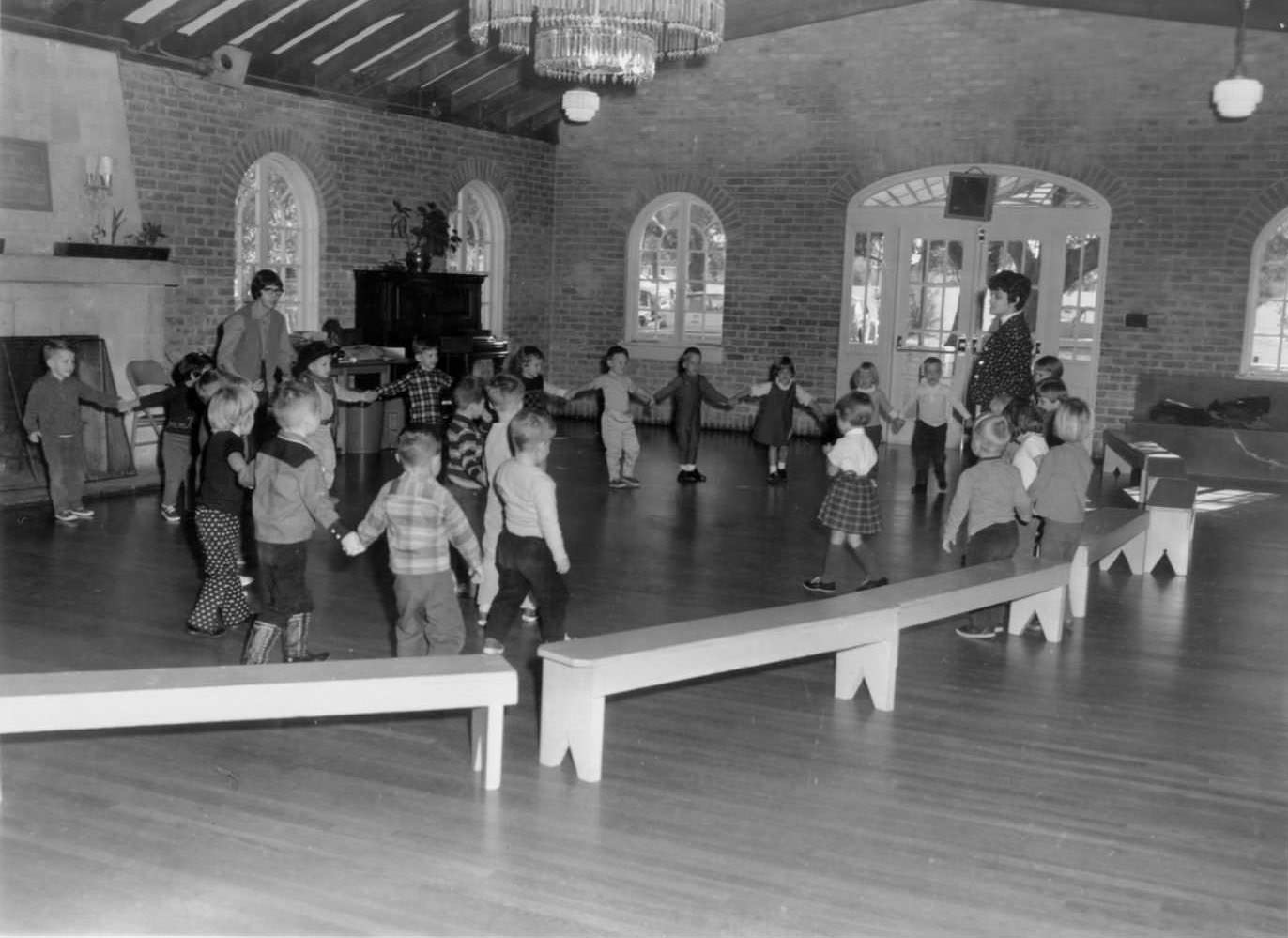 A group of children playing a game. They are in a large circle, holding hands, with two adult instructors, 1966