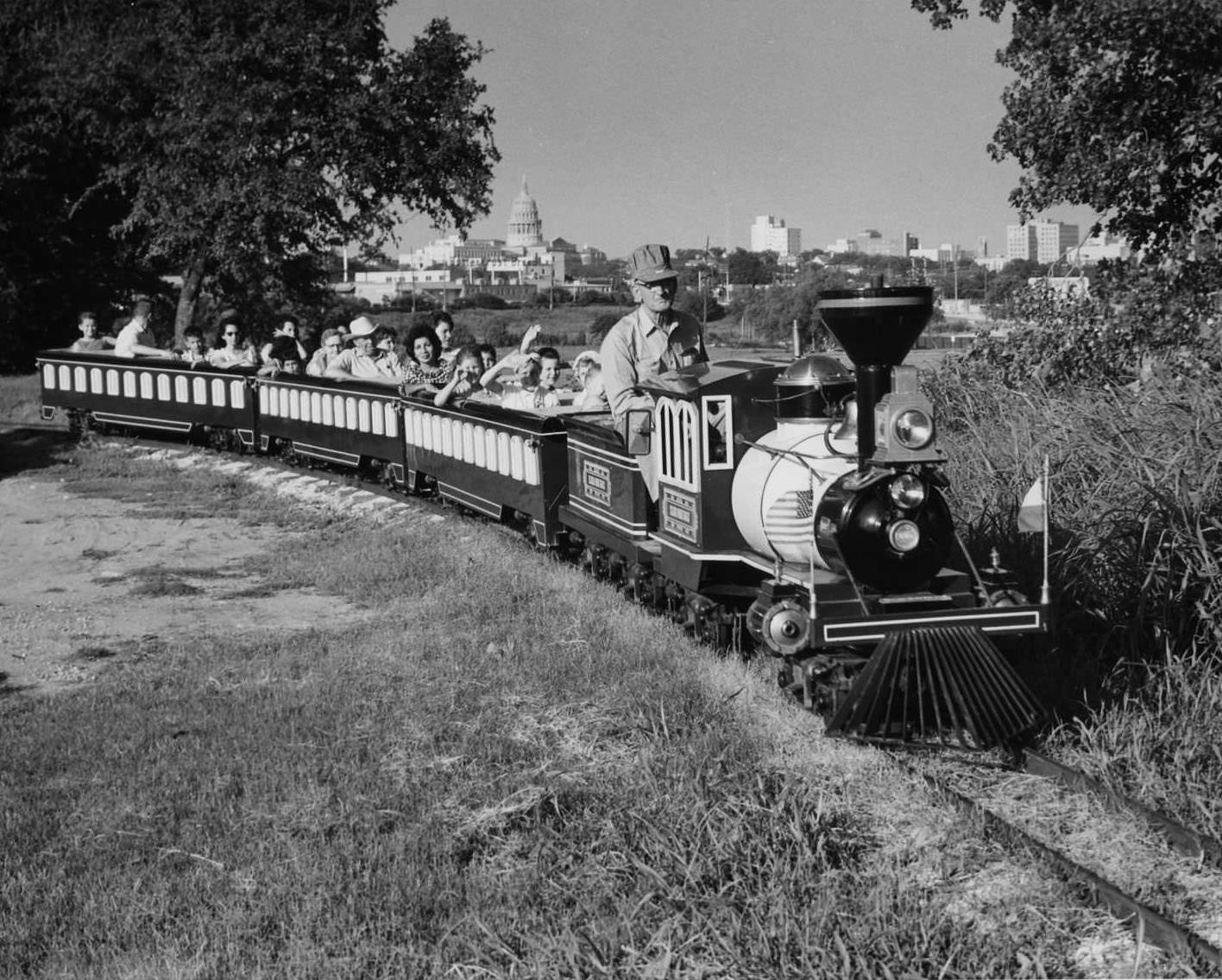 Children and parents riding the Zilker Zephyr miniature train through Zilker Park. Downtown Austin and the Capitol building are visible in the background, 1960s