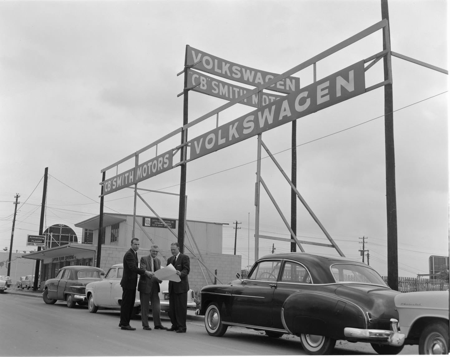 The exterior of CB Smith Volkswagen Motors, Austin, 1960. There are a few cars parked next to the sign advertisement. All three of the men are wearing suits.