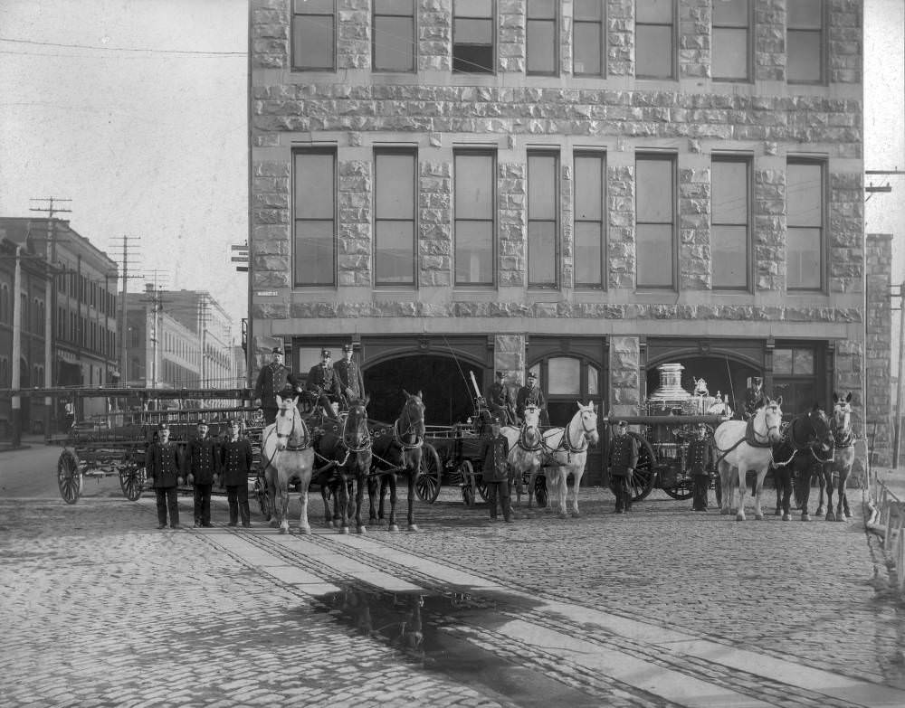Firemen stand and pose in front of the Denver Fire Department Headquarters with Hook & Ladder No. 1 and Hose No. 4 in the first floor of the City Hall Building at 14th (Fourteenth) and Market Street in downtown Denver, 1900