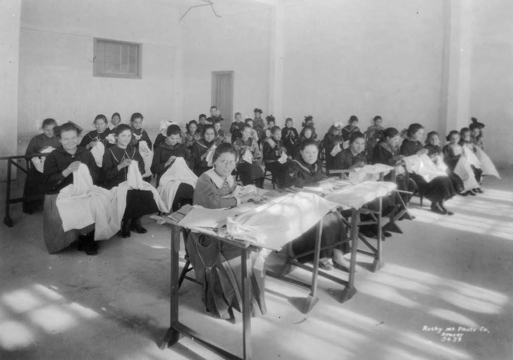 Girls and their teacher sit at tables and on chairs and sew in a classroom at the Denver Orphans' Home (later the Denver Children's Home) at 1501 Albion Street in the South Park Hill neighborhood of Denver, 1909