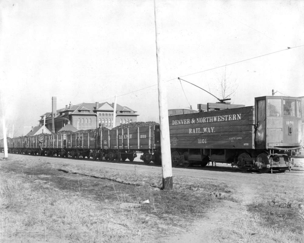 View of Leyden Mine coal trains, Denver & Northwestern Railway cars number 1101, 1555, 1523, and 1546 with Alcott Public School at West 41st (Forty-first) Avenue and Tennyson Street, Denver, 1900