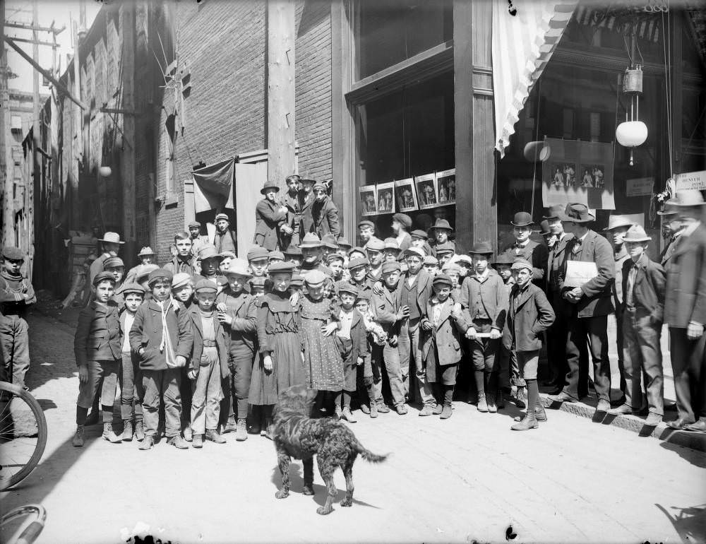 A group of children, probably delivery boys and girls, pose outside of the Denver Post newspaper office on 16th (Sixteenth) Street in Denver, 1900