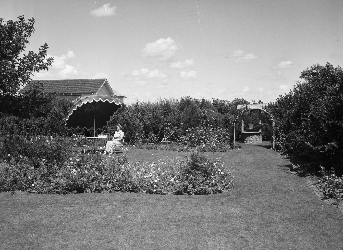 Mrs. C. H. Bertram, shown sitting at an umbrella-protected table in her "outdoor living room," which is surrounded with flowerbeds and hedges, 1940
