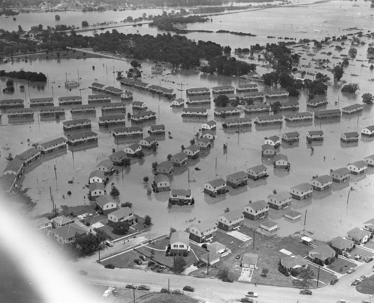 Fort Worth, Texas, flood of 1949, showing a flooded neighborhood. There are few cars on the roads that have not been completely flooded. Groups of people are gathered in front of their homes and in the streets.