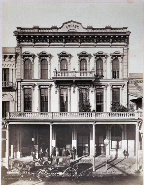 View of the three-story Arcade Hotel with balcony across the second floor, partial balcony at third floor, 1875