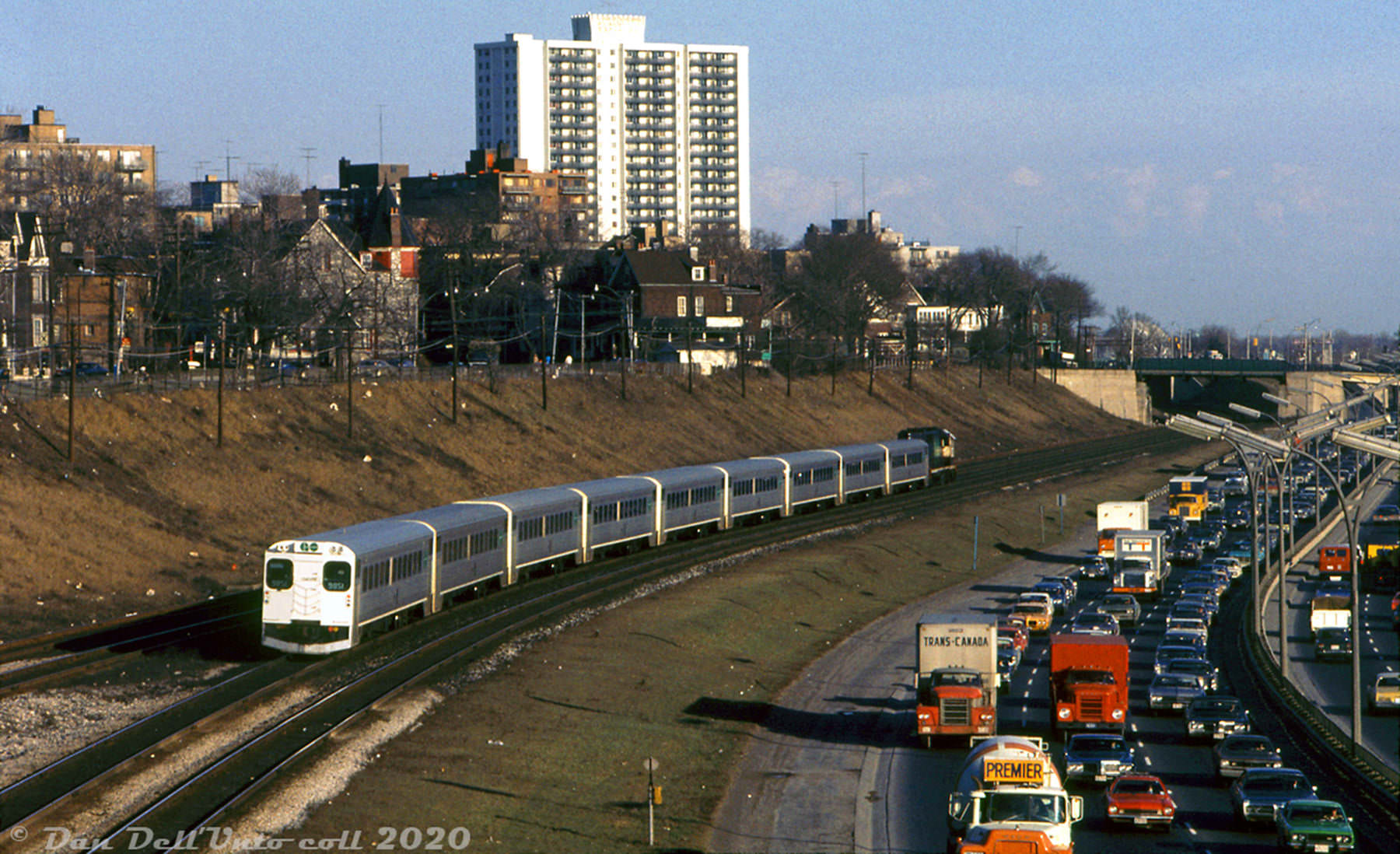 A grungy March 1973 view along the Gardiner Expressway at Sunnyside, showing an inbound GO train heading to Union Station as PM rush hour traffic inches along the Gardiner out of the city.