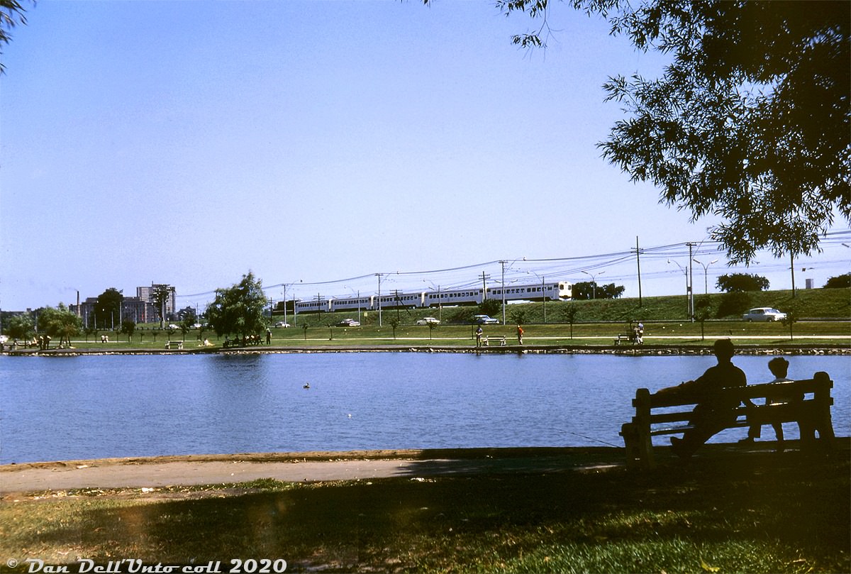 Enjoying a Summer's day in 1967 around Grenadier Pond, onlookers pay little attention to a passing "GO train" on the Lakeshore line - at the time a new experimental commuter train service operated by the Government of Ontario, that eventually grew into the present day GO Transit system.
