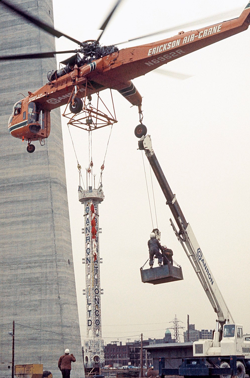 Bob Benns (seen on the ground) managing the last antenna piece lift by Olga the Sikorsky helicopter, 1975