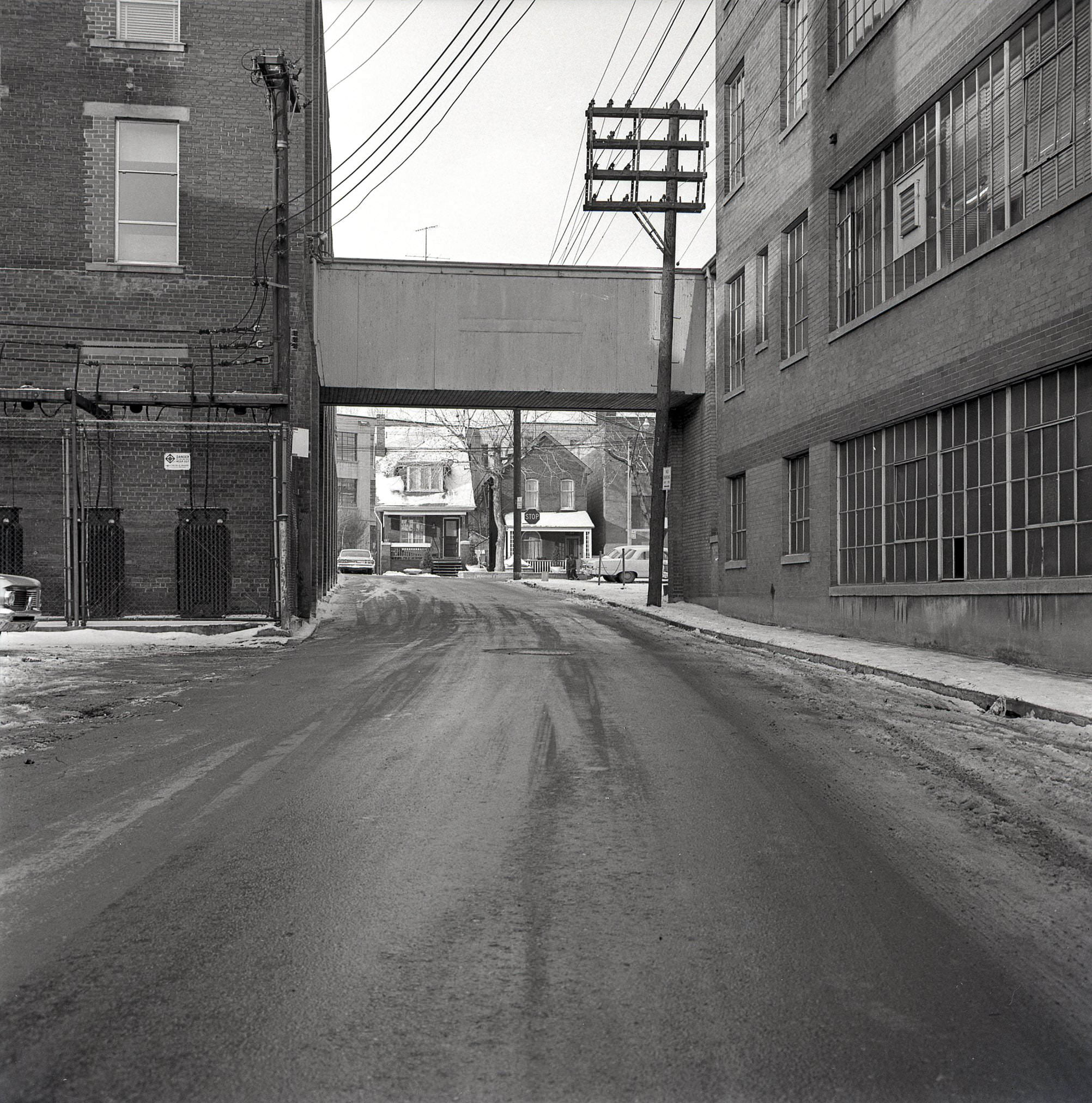 Above-ground connection between two buildings at the west end of Pears Avenue, framing a view of a couple houses on Bedford Road, 1964