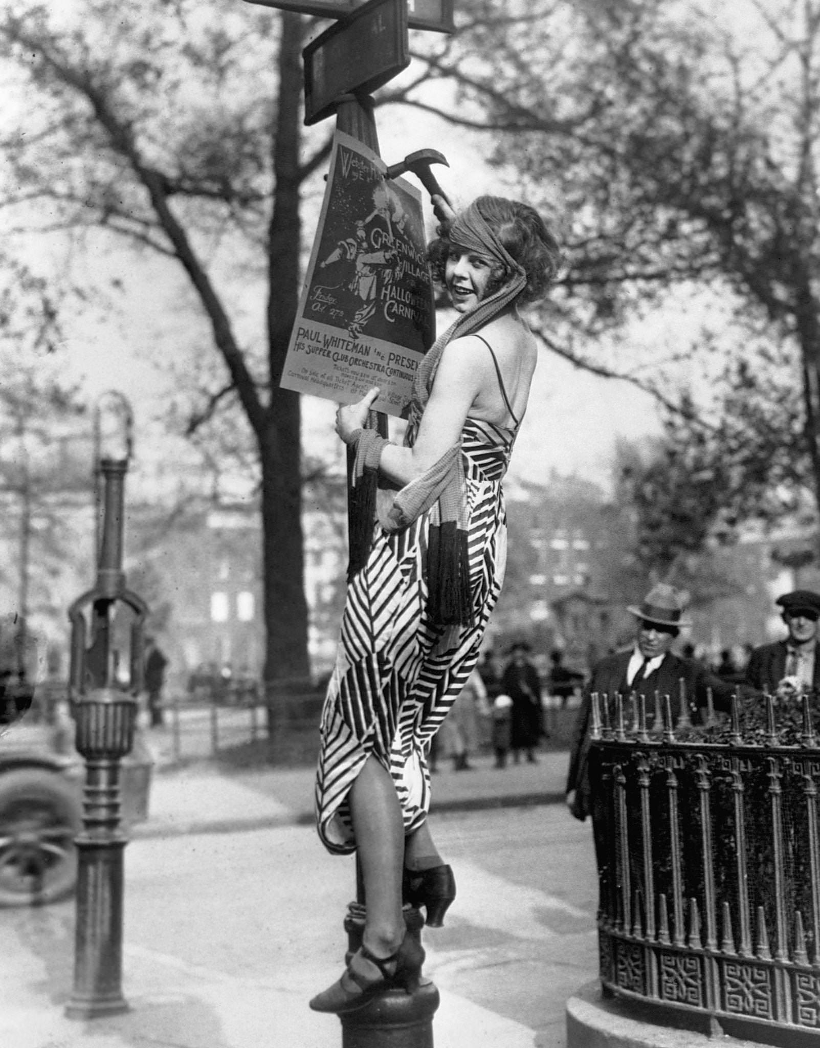 A flapper hangs a poster to advertise the Greenwich Village Halloween Ball, at which Paul Whiteman is to perform.