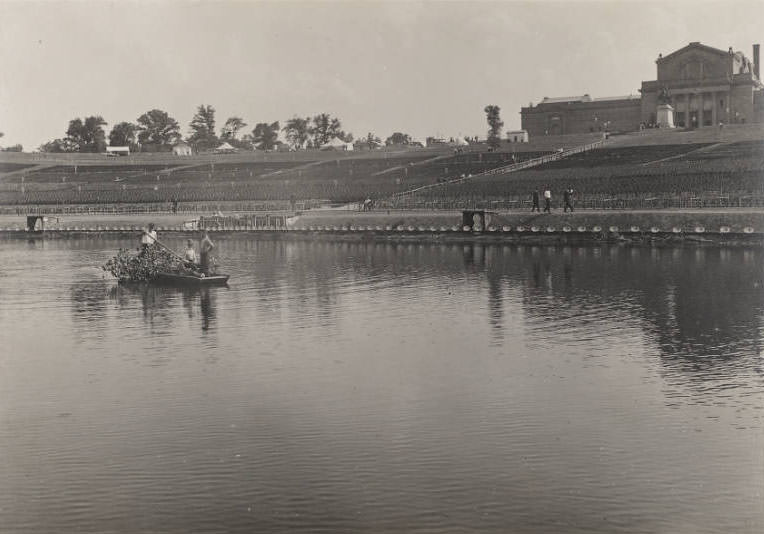 Three men in a row boat clear debris from the Grand Basin, Forest Park, in preparation for performances of the Pageant and Masque of St. Louis, 1914