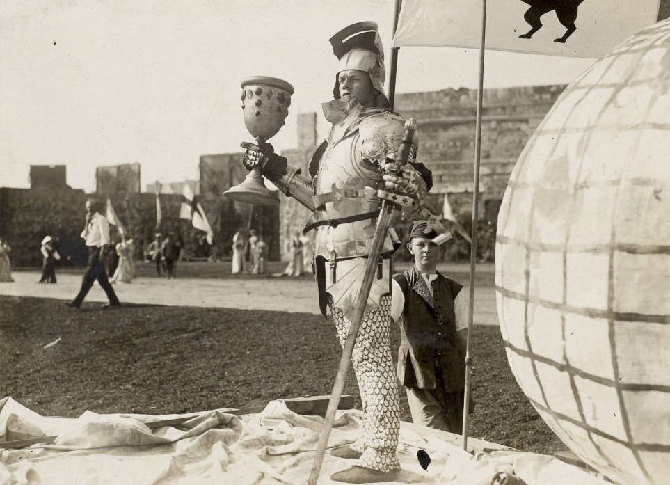 Actor holding a huge goblet portrays a French knight in armor in the Masque, Pageant and Masque of St. Louis. A young man in costume attends him.