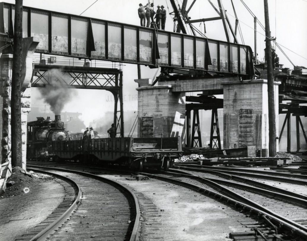 Men setting a girder for elevated railway. Below, a locomotive is in the process of switching cars, 1910