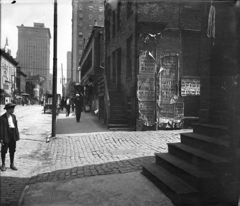A boy in the foreground is reading an assortment of advertisements plastered to the side of a brick building, 1910