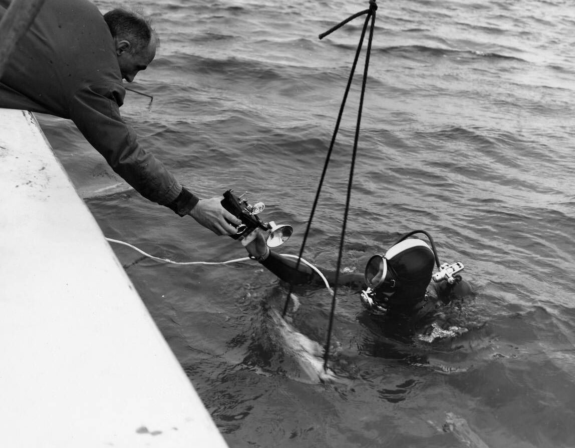 A man on a boat handing a camera to a scuba diver, the two men were working as part of a joint effort between the Army Corps of Engineers and the State of Alaska to study tides and surf action on the Homer Spit,