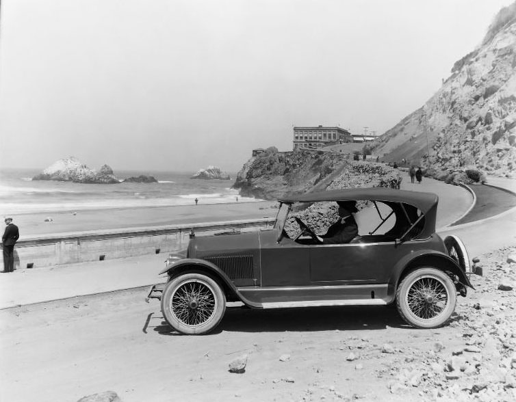 Historic San Francisco Cliff House. Road and sidewalk construction has been completed on the curved road including new landscape work. A military man in uniform and Seal Rocks can be seen along the left, 1922