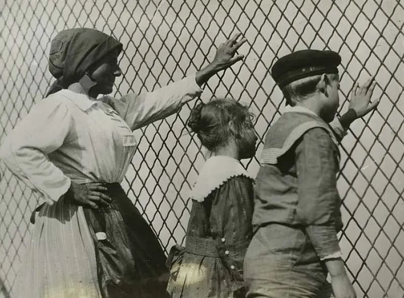 A woman, a boy and a girl at a chain link fence, Ellis Island, New York